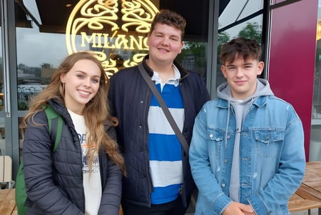 Drei Schüler der Newtown School stehen vor einem Café mit einem dekorativen Schild im Hintergrund.