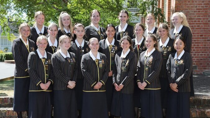 Schülerinnen der Nga Tawa Diocesan School stehen in schwarzen und goldenen Roben im Freien vor einem Backsteingebäude.