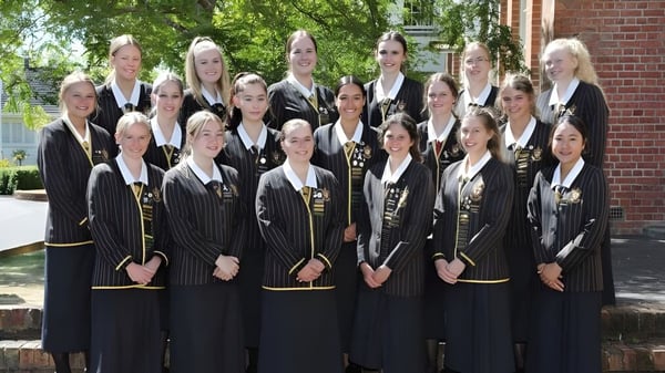Eine Gruppe junger Schülerinnen in schwarzen akademischen Roben steht auf dem Campus der Nga Tawa Diocesan School vor einem Backsteingebäude im Grünen.