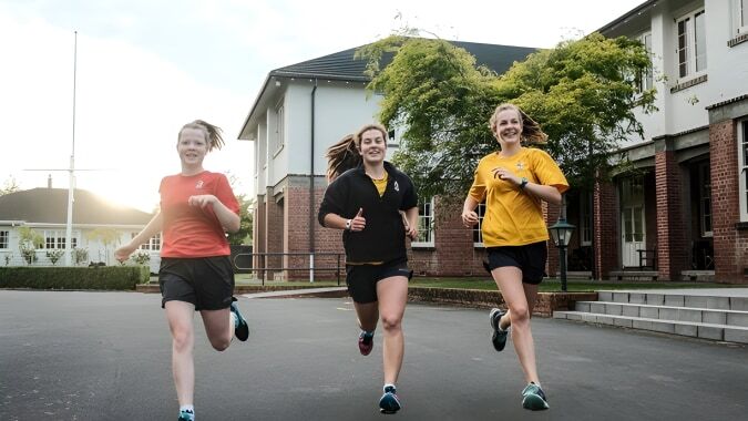 Drei Schülerinnen der Nga Tawa Diocesan School joggen gemeinsam auf einem gepflasterten Weg vor einem Backsteingebäude.