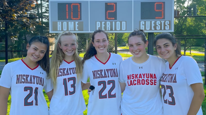 Eine Gruppe von fünf Schülerinnen der Niskayuna High School steht vor einer Anzeigetafel während eines Lacrosse-Spiels auf dem Sportplatz.