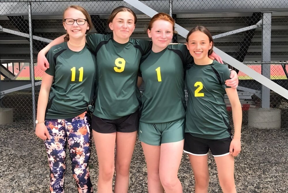 Vier Schülerinnen der Noble Central School stehen gemeinsam in grünen Sporttrikots vor einem Maschendrahtzaun.