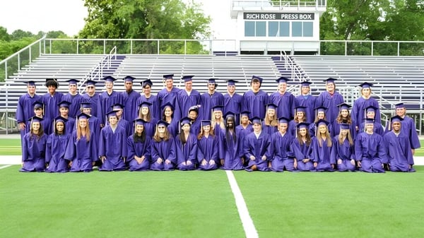 Eine Gruppe von Schülerinnen und Schülern der Norfolk Christian Schools steht in violetten Abschlussroben auf einem Feld vor einer stadionähnlichen Struktur.