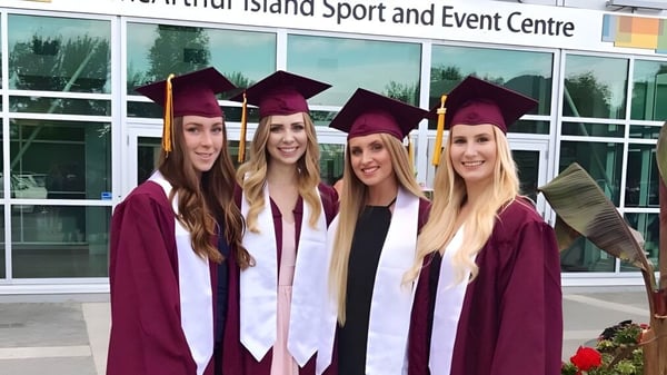 Vier Schülerinnen der NorKam Secondary School stehen in Abschlussgewändern vor dem Whidbey Island Sport and Event Centre.