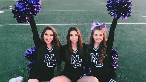 Drei Cheerleader der North Canyon High School stehen auf einem grünen Feld und halten lila und schwarze Pompons.