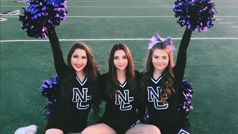 Drei Cheerleader der North Canyon High School stehen auf einem grünen Feld und halten lila und schwarze Pompons.