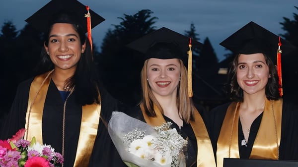 Drei Absolventen der North Delta Secondary School in Talaren stehen gemeinsam vor Bäumen und dunklem Himmel.