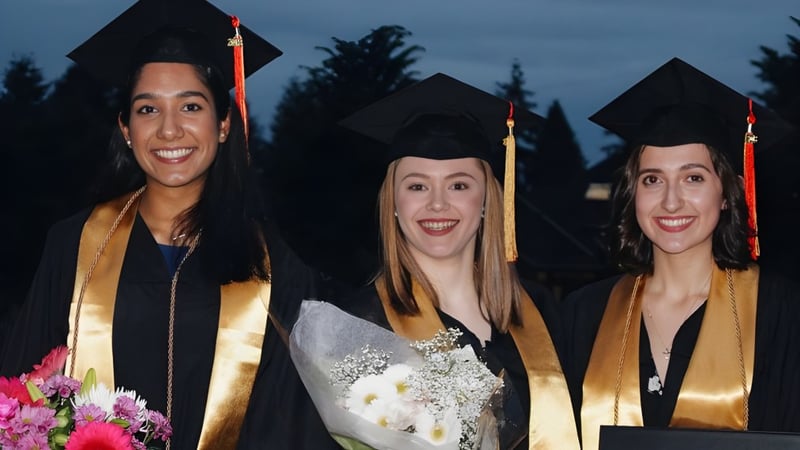 Drei Absolventen der North Delta Secondary School in Talaren stehen gemeinsam vor Bäumen und dunklem Himmel.