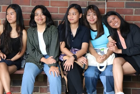 Fünf Schülerinnen mit langen dunklen Haaren sitzen zusammen auf einer Mauer am North Lake Senior Campus und lächeln in die Kamera.
