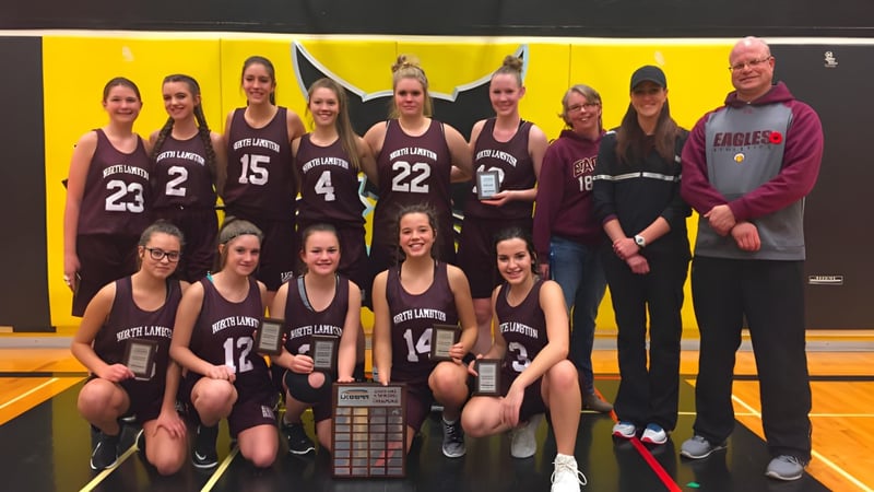 Eine Gruppe junger Basketballspielerinnen posiert mit einem Pokal vor einem gelben Hintergrund auf dem Campus der North Lambton Secondary School.
