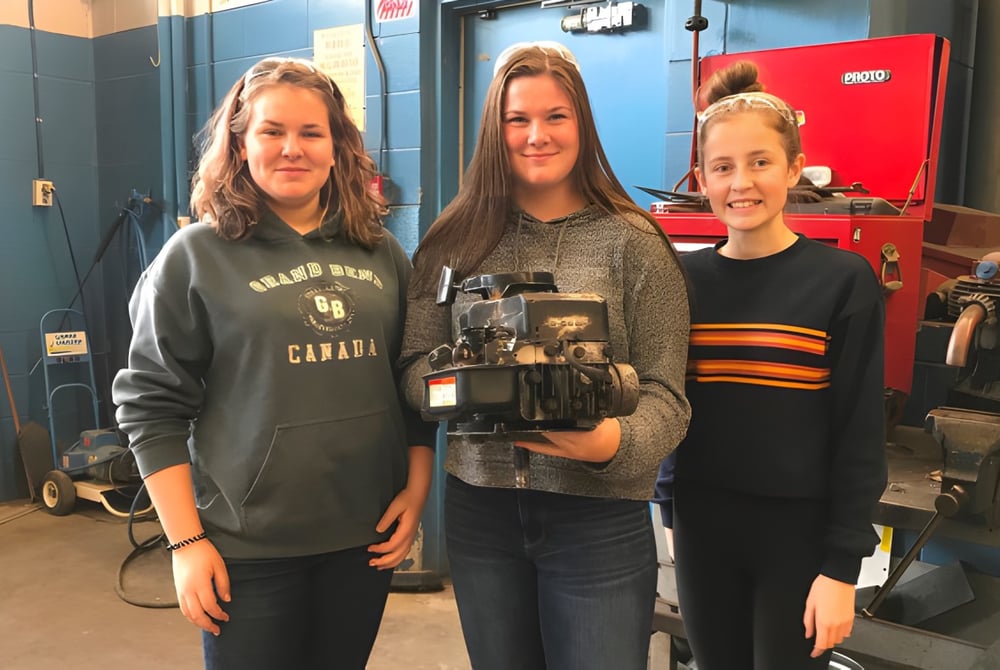 Drei Schülerinnen stehen gemeinsam in einer Werkstatt der North Lambton Secondary School mit Werkzeugen im Hintergrund.