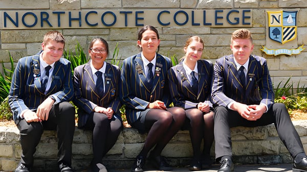 Eine Gruppe von fünf Schülern in Schuluniform sitzt auf einer Bank vor einem Schild des Northcote College.