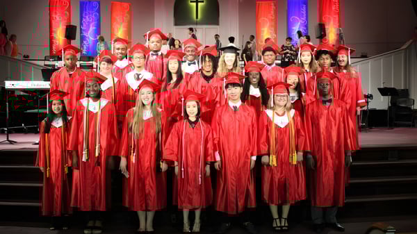 Eine Gruppe von Absolventinnen und Absolventen der Northside Christian Academy steht in roten Talaren in einer Kirche mit Kreuz im Hintergrund.