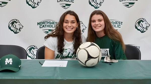 Zwei Schülerinnen der Northwest Catholic High School sitzen an einem Tisch mit Rugbyball vor einem Logo-Hintergrund.
