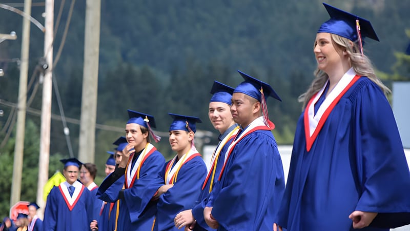 Eine Gruppe von Absolventen der Norwell District Secondary School steht zusammen in blauen Abschlussroben vor einem bewaldeten Hintergrund.