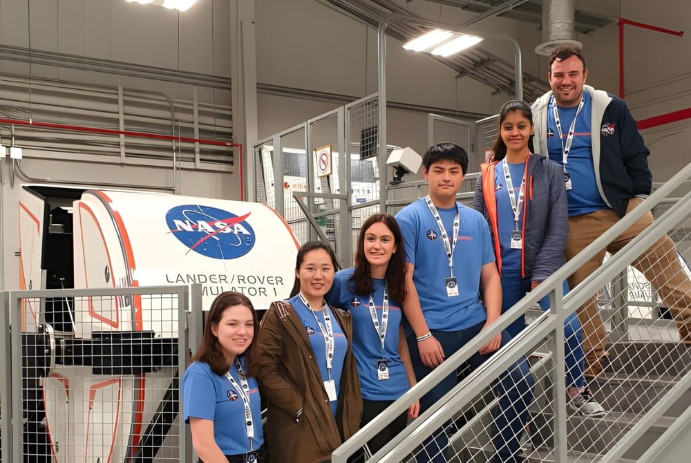 Eine Gruppe von Schülerinnen und Schülern der Norwood International High School steht in NASA-Uniformen vor einem großen NASA-Logo und einem Lander Rover Modell.