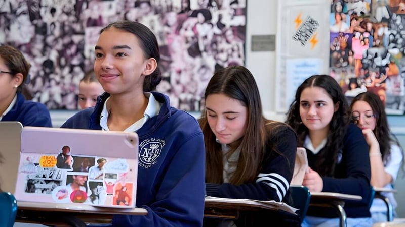Eine Gruppe von Schülern sitzt im Klassenzimmer der Notre Dame High School mit einer Collage aus Bildern und Postern im Hintergrund.