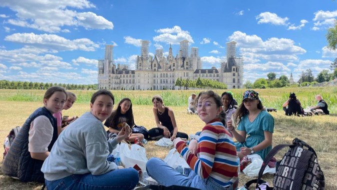 Eine Gruppe von Menschen befindet sich auf einer Wiese vor dem Schlossähnlichen Gebäude der Notre Dame Saint Joseph Schule unter blauem Himmel.