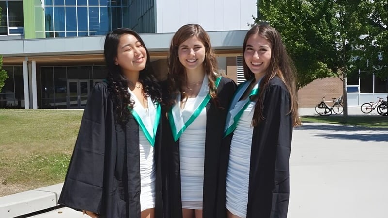 Drei Schülerinnen der Oak Bay Secondary High School stehen in Abschlussroben vor einem Universitätsgebäude mit Fahrrädern und Grünfläche.