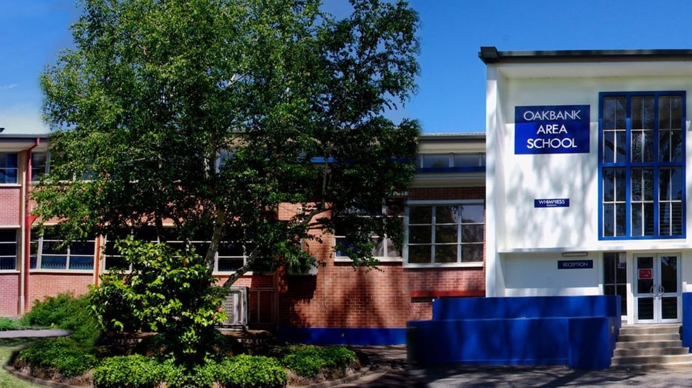 Das zweistöckige Backsteingebäude der Oakbank Area School mit einem blauen Schild für den Betreuungsbereich und grüner Umgebung ist zu sehen.