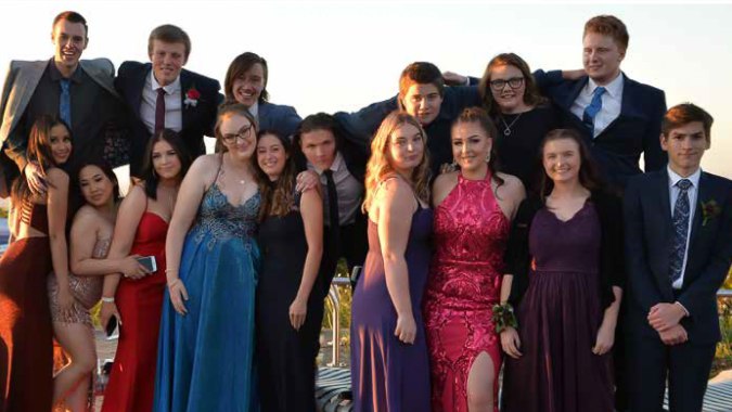 Eine Gruppe von Personen in formeller Kleidung steht im Freien vor einem Gewässer und Sonnenuntergang auf dem Campus der Oakbank Area School.