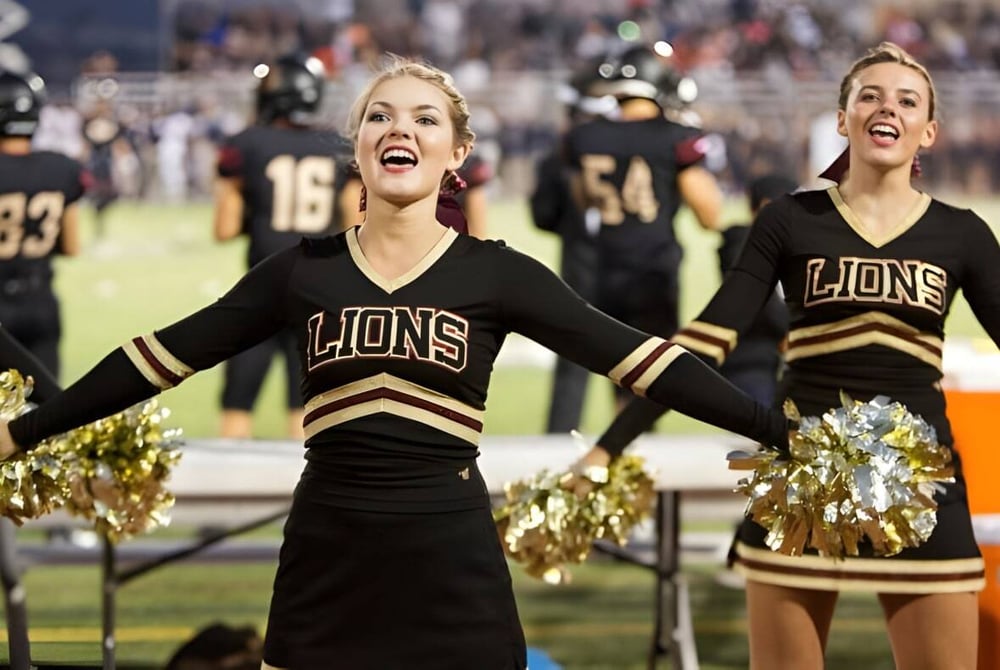 Zwei Cheerleader der Oaks Christian School in schwarz-goldenen Uniformen feuern am Spielfeldrand eines Footballspiels an.