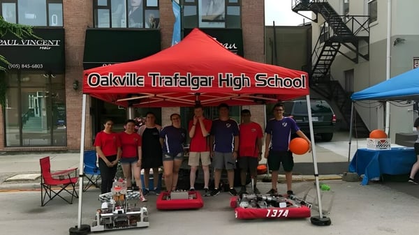 Eine Gruppe Schüler der Oakville Trafalgar High School steht vor einem roten Zelt mit dem Schulnamen neben einem Roboter und Ausrüstung auf dem Boden.
