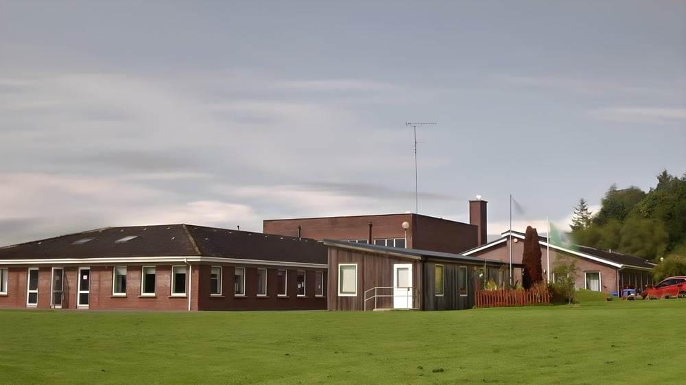 Das Schulgebäude des O’Carolan College steht auf einer Wiese und ist von Bäumen umgeben.