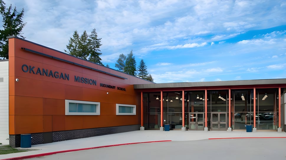 Das moderne orangefarbene Gebäude der Okanagan Mission Secondary School mit großen Fenstern und Kiefern im Umfeld.
