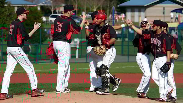 Eine Gruppe Baseballspieler des Olympic College feiert auf dem Spielfeld mit Gebäude und Bäumen im Hintergrund.