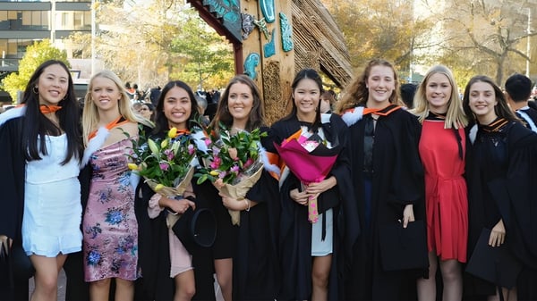 Eine Gruppe Absolventinnen der Onehunga High School steht vor einem farbenfrohen Wandbild und hält Blumensträuße.
