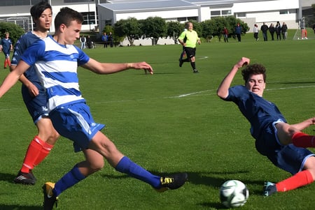 Zwei Fußballspieler der Onehunga High School kämpfen auf dem Spielfeld um den Ball, während weitere Spieler und ein Schiedsrichter im Hintergrund zu sehen sind.