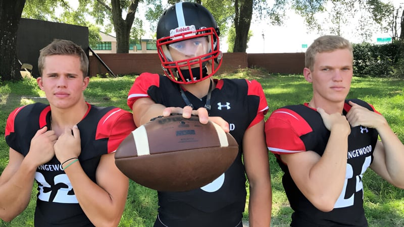 Drei Schüler der Orangewood Christian School in Sportuniformen stehen mit einem Football in einem parkähnlichen Gebiet.