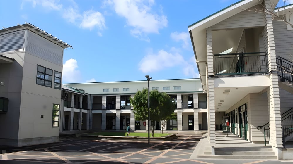 Das mehrstöckige Hauptgebäude des Orewa College mit Innenhof und Wegen, umgeben von Bäumen unter blauem Himmel.