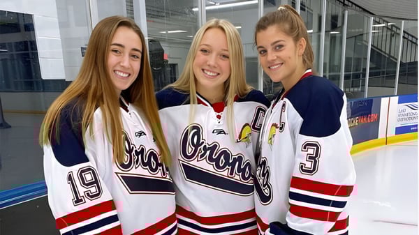 Drei Schülerinnen der Orono High School stehen in Hockey-Jerseys auf der Eisbahn in der Sporthalle.