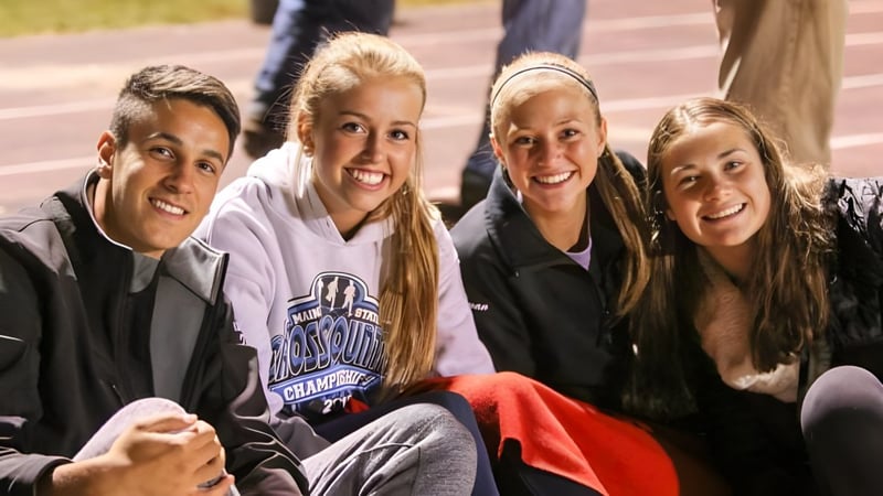 Eine Gruppe von vier Schülern der Orono High School sitzt lachend auf einem Sportplatz vor einem Backsteingebäude.