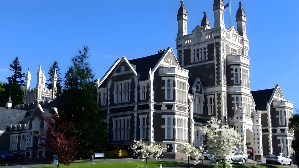 Das gotische Hauptgebäude der Otago Boys' High School ist von blühenden Bäumen vor blauem Himmel umgeben.