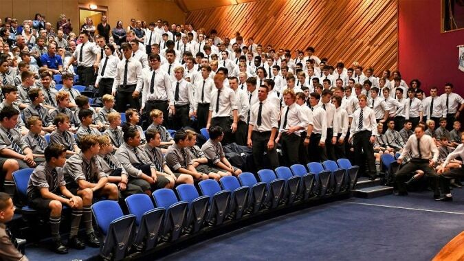 Eine große Gruppe Schüler der Otago Boys' High School sitzt in weißen Uniformen in einem auditorium mit holzvertäfelten Wänden.