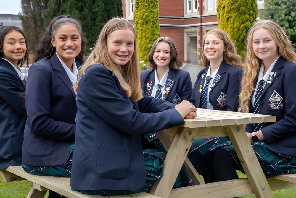 Eine Gruppe Schülerinnen der Otago Girls High School sitzt auf einer Holzbank im Gartenbereich mit Bäumen und Schulgebäuden im Hintergrund.