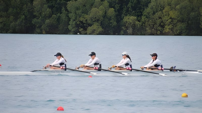 Vier Schülerinnen der Otago Girls High School rudern auf einem ruhigen See, umgeben von einem bewaldeten Landschaftshintergrund.
