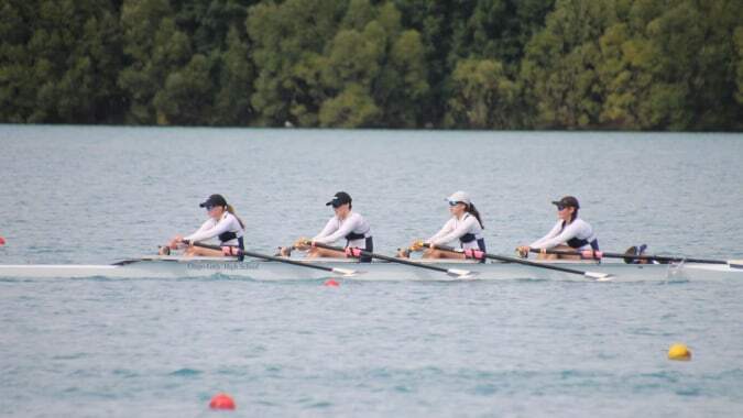 Schülerinnen der Otago Girls High School beim Rudern auf einem See inmitten einer bewaldeten Landschaft.