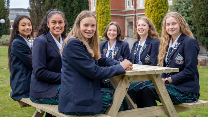 Eine Gruppe von Schülerinnen der Otago Girls High School sitzt zusammen auf einer Holzbank im grünen Außenbereich.