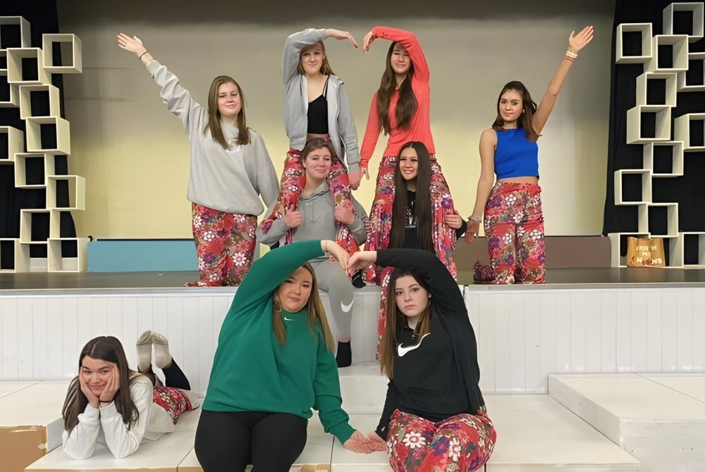 Eine Gruppe junger Schülerinnen der Our Lady Catholic College steht in bunten Outfits auf einer Bühne mit geometrischem Hintergrund.