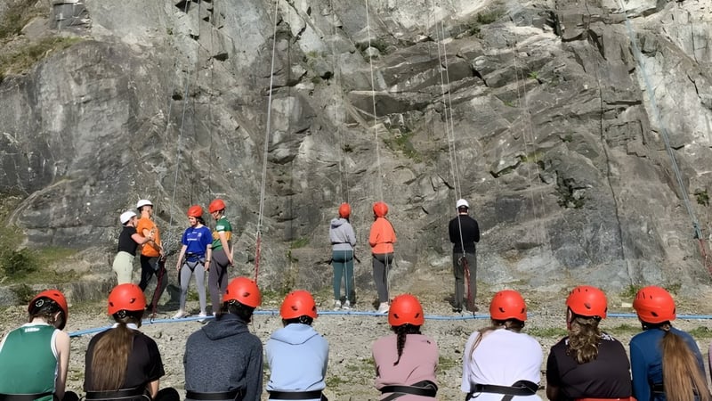 Eine Gruppe Schüler in bunten Jacken steht vor einer großen, zerklüfteten Felsenformation auf dem Gelände von Our Lady's College.
