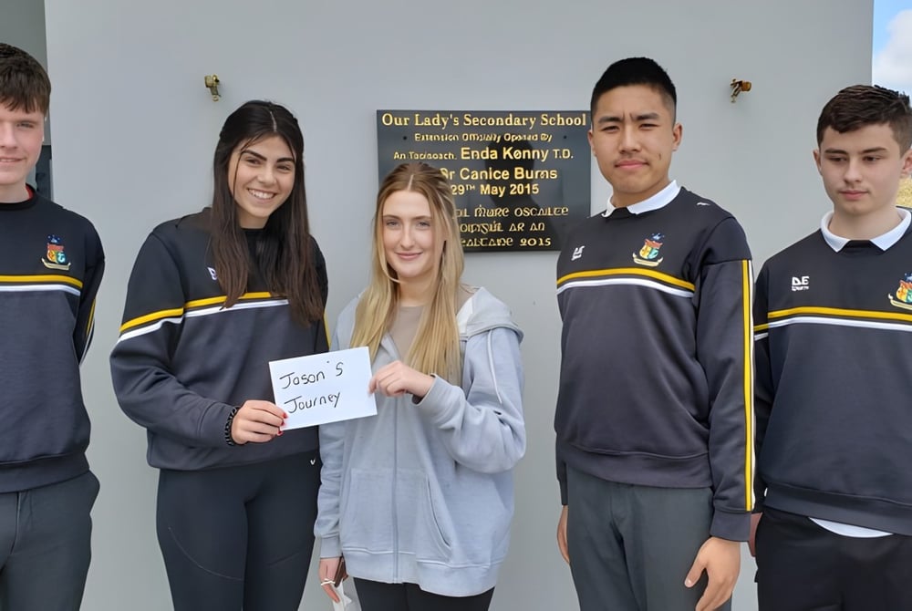 Eine Gruppe Schüler in Schuluniformen steht vor einem Schild der Our Lady’s Secondary School, während ein Schüler ein weiteres Schild hält.