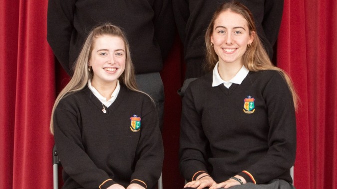 Zwei Schülerinnen der Our Lady’s Secondary School stehen vor einem roten Vorhang und lächeln in die Kamera.