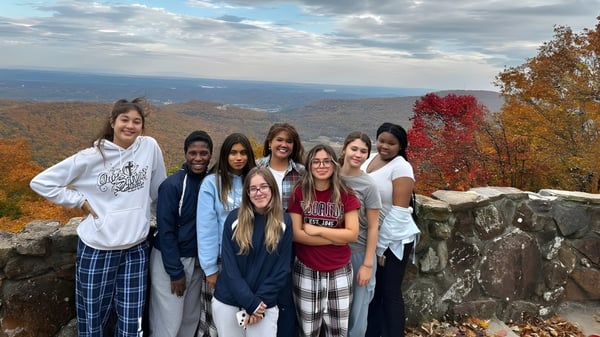 Eine Gruppe Schülerinnen und Schüler der Our Savior New American School steht auf einem Felsen mit Blick auf eine bunte Herbstlandschaft.