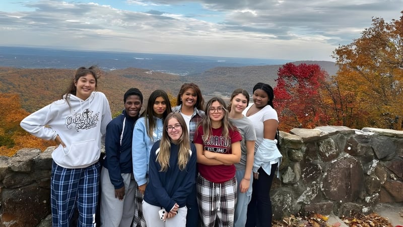 Eine Gruppe Schülerinnen und Schüler der Our Savior New American School steht auf einem Felsen mit Blick auf eine bunte Herbstlandschaft.