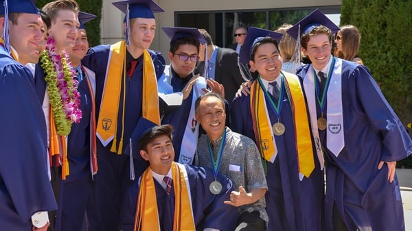 Absolventen der Pacifica Christian High School stehen in lila und goldener Abschlusskleidung zusammen bei der Graduierungsfeier.