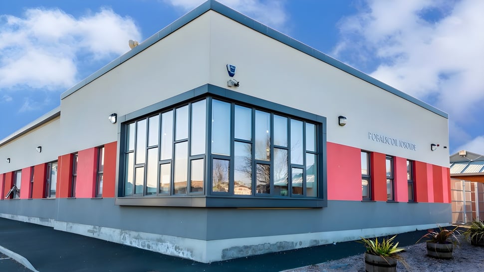Das moderne Gebäude der Palmerstown Community School mit großen Glasfenstern vor blauem Himmel und weißen Wolken.